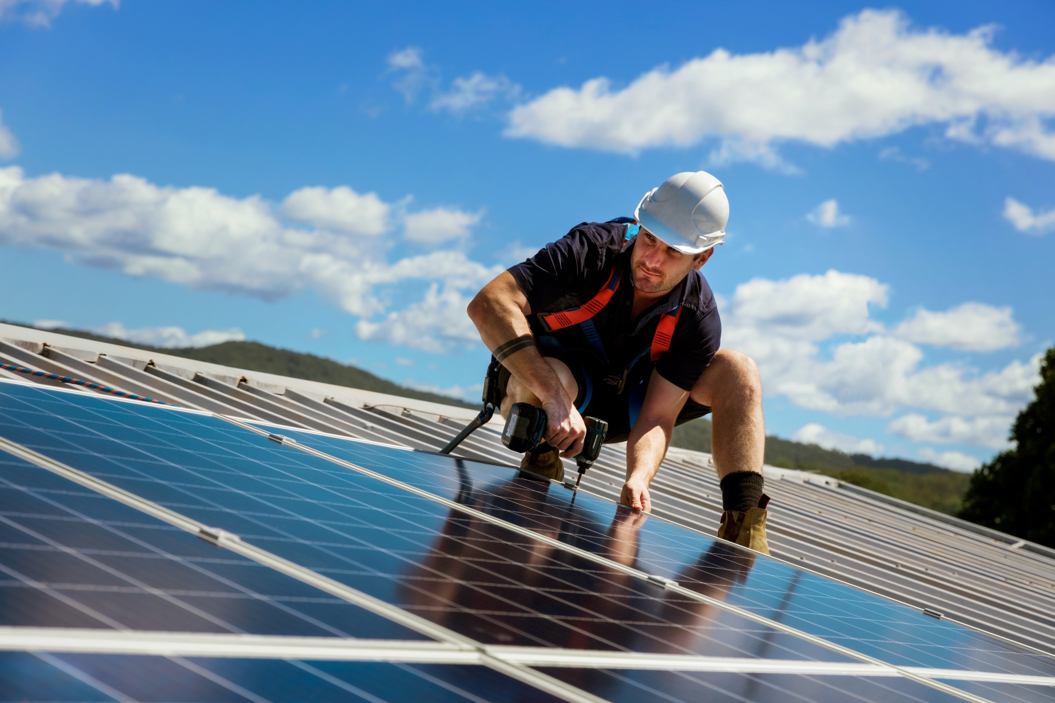 Ein Arbeiter mit weissem Schutzhelm und Sicherheitsgeschirr montiert Solarmodule auf einem Metalldach unter blauem Himmel.