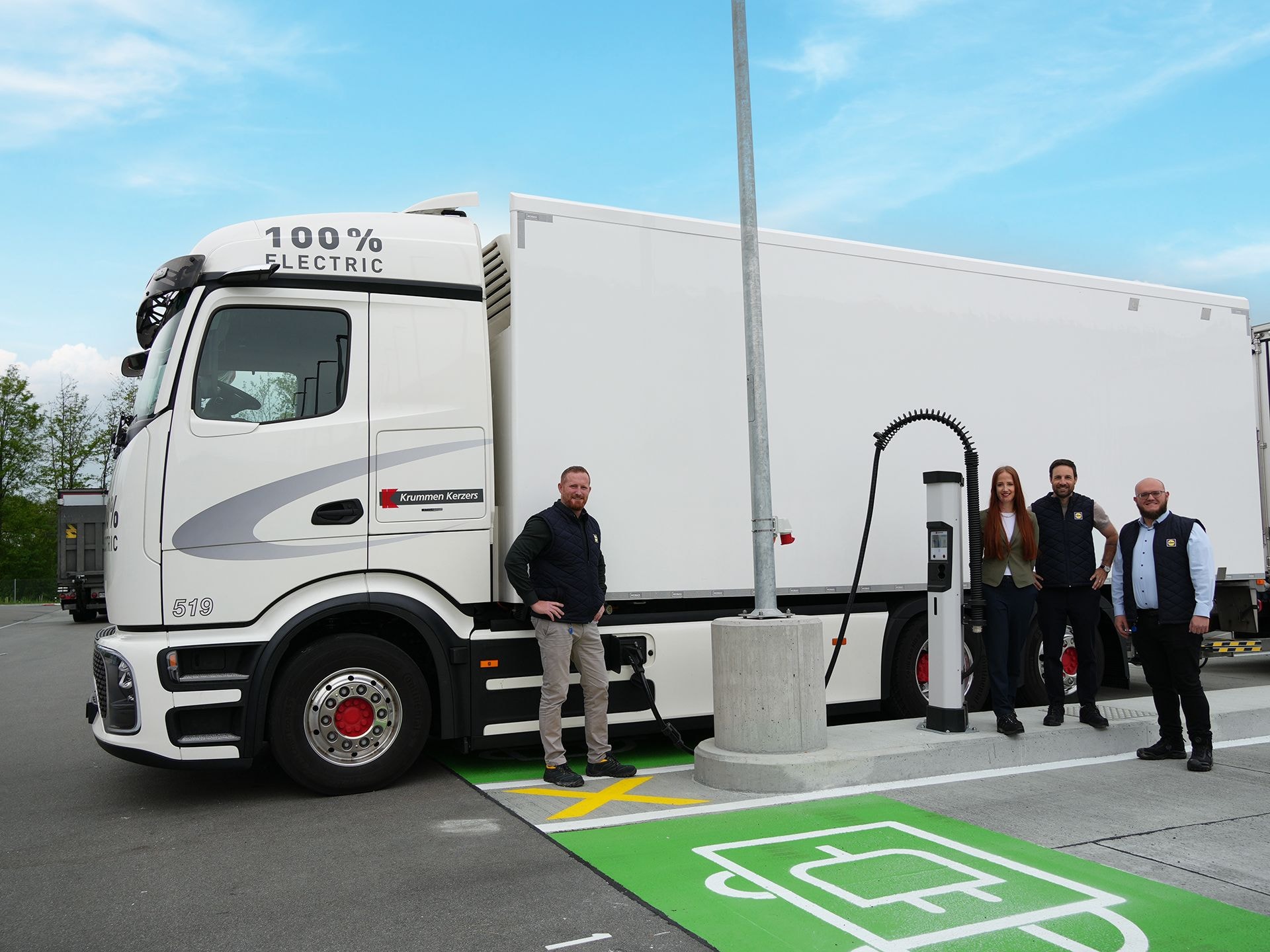 Grosser weisser Lkw steht auf einem elektropakplatz, davor stehen drei Personen, die freundlich lächeln