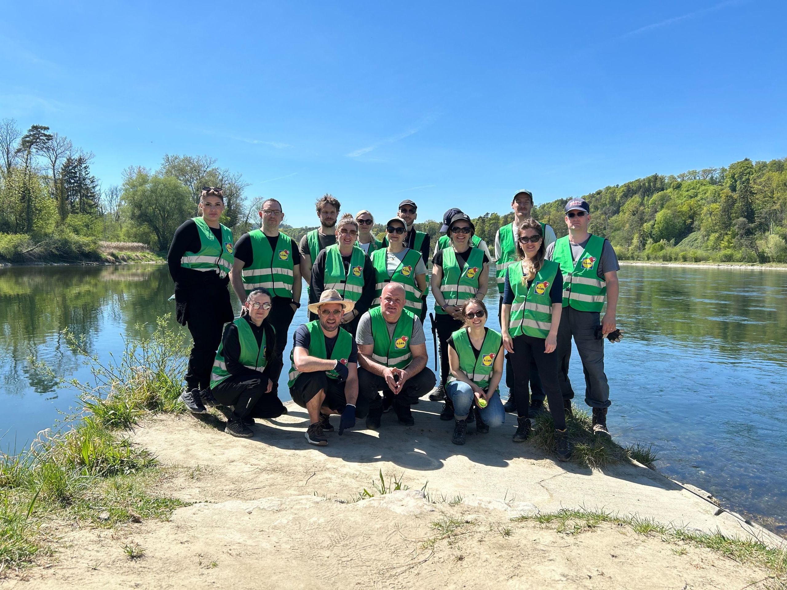 Elf Lidl-Schweiz-Mitarbeitende in grünen Warnwesten posieren in zwei Reihen auf einer Sandbank an einem naturnahen Fluss – Teamfoto nach dem gemeinsamen Einsatz zur Aufwertung wertvoller Lebensräume für die Gelbbauchunke und weitere geschützte Arten in der Schweiz.
